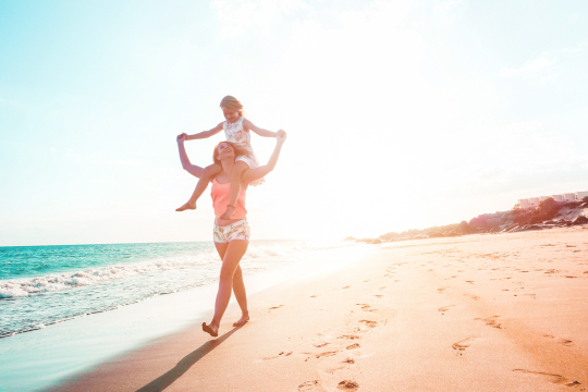 Maedchen auf den Schultern ihrer Mutter, spazieren im Sonnenuntergang am Meer entlang
